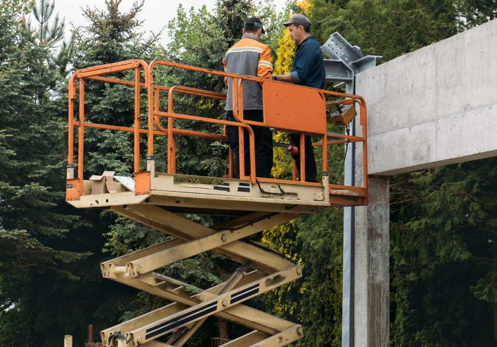 Two workers in safety gear operating orange scissor lift near concrete bridge structure"- Scissor Lift Training