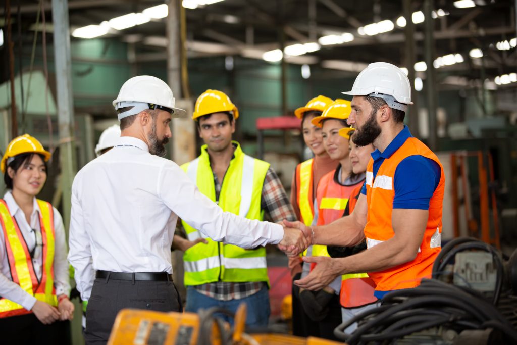 Construction manager shaking hands with worker while team in safety gear watches in warehouse