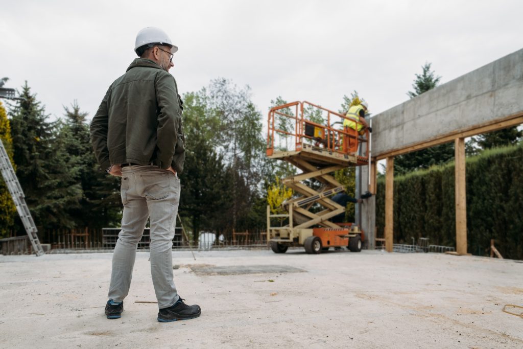 Construction supervisor in hard hat observing orange scissor lift at concrete building sit